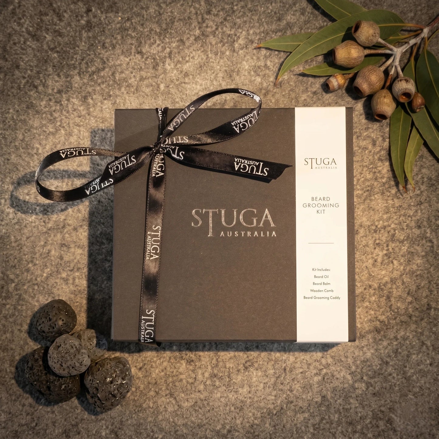 Stuga Beard Grooming Kit in a matte black gift box tied with a Stuga-branded satin ribbon, with eucalyptus leaves and gum nuts on a dark stone background.