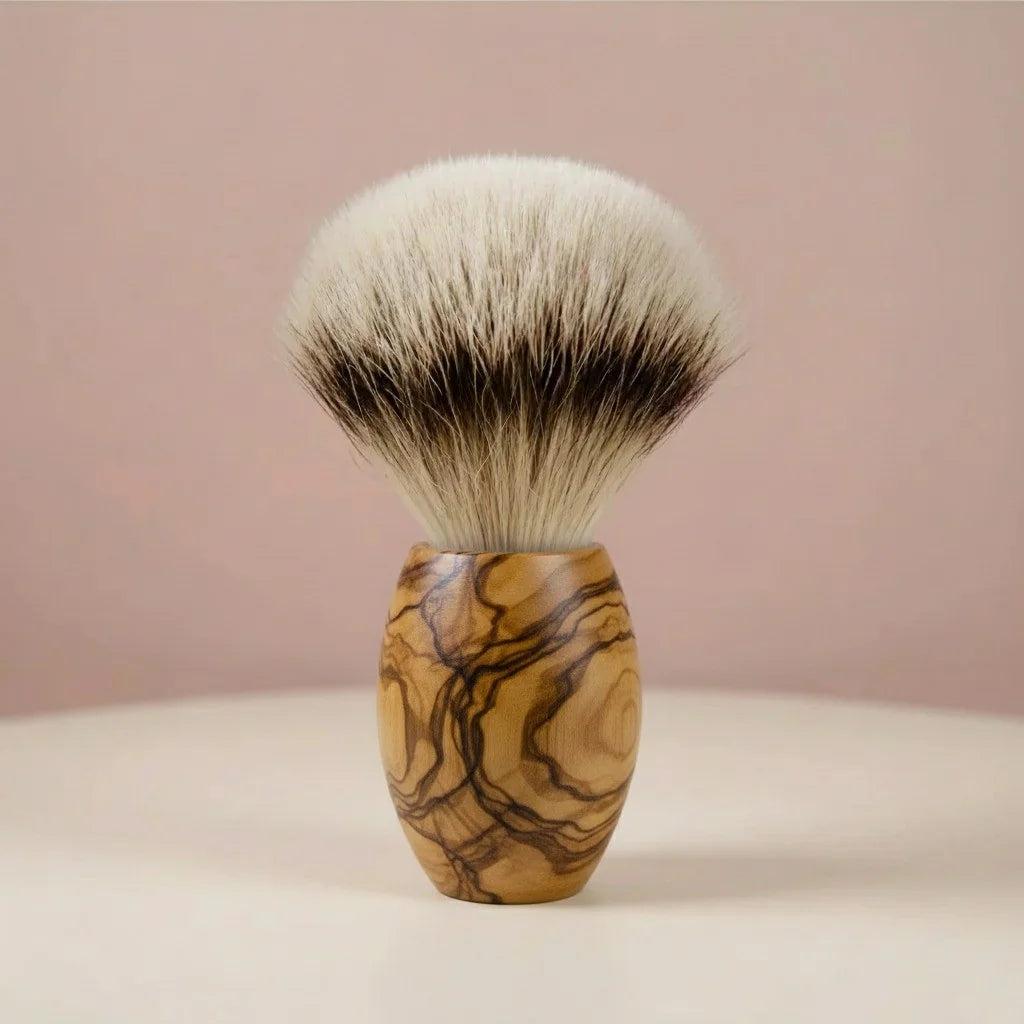Shaving brush with a natural olive wood handle and soft, silver-tipped badger bristles, standing upright against a pink backg