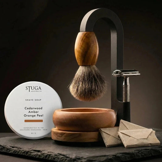 Premium Magic Shaving Kit — badger brush with olive wood handle suspended from black arch stand, STUGA shave soap tin, handturned timber bowl, safety razor and wrapped blades on slate