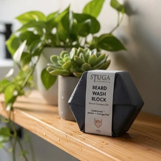Stuga beard wash block in dark grey packaging with white label, on a wooden shelf with green plants.