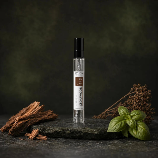 Stuga Cedarwood No. 443 cedarwood perfume, 10ml eau de parfum spray bottle on dark stone with cedarwood shavings, fresh basil leaves and dried botanicals.