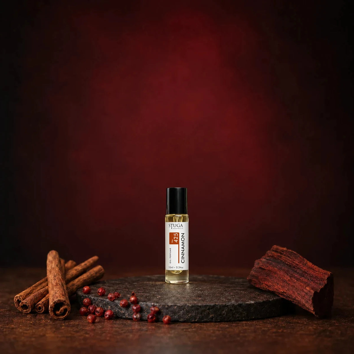 Stuga Cinnamon No. 425 cinnamon oil perfume, 10ml roll-on bottle on a dark stone disc with cinnamon sticks, pink peppercorns and mahogany wood against a deep red backdrop.