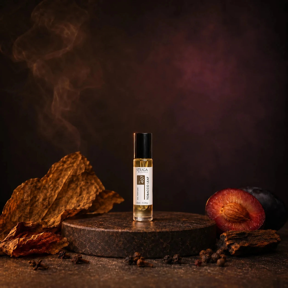 Stuga Tobacco Leaf No. 407 tobacco oil perfume, 10ml roll-on bottle on a dark stone plinth with dried tobacco leaves, a spiced plum and drifting smoke.