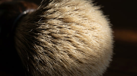 Close-up of a luxury silvertip badger shaving brush knot with soft pale tips and a hand-turned timber handle, warm light on a dark surface