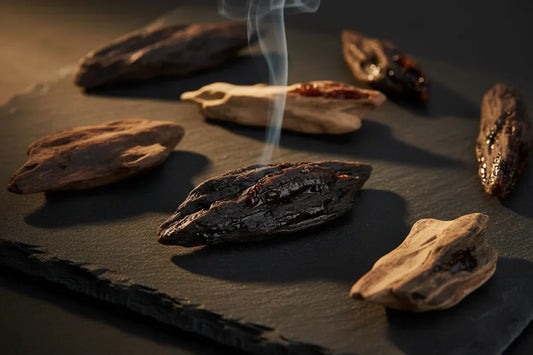 Dark oud agarwood chips with resin deposits on black stone with a wisp of smoke rising