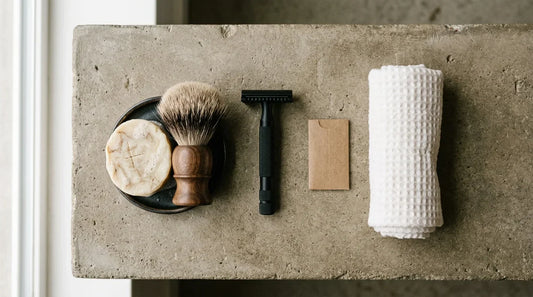 Beginner safety razor kit flatlay - matte black double-edge razor, badger brush, artisan soap puck, blade pack and rolled white towel on warm grey concrete