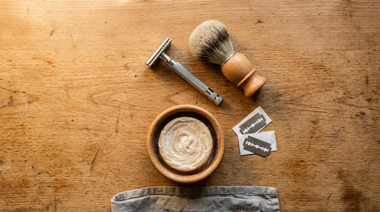 Traditional wet shaving kit flat lay on warm timber — safety razor, badger brush, shaving bowl with soap, blades and linen cloth