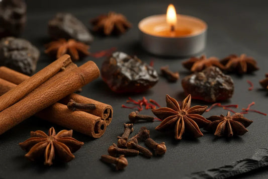 Winter spices and perfume ingredients — cinnamon, cloves, star anise, amber resin, and oud chips with candlelight