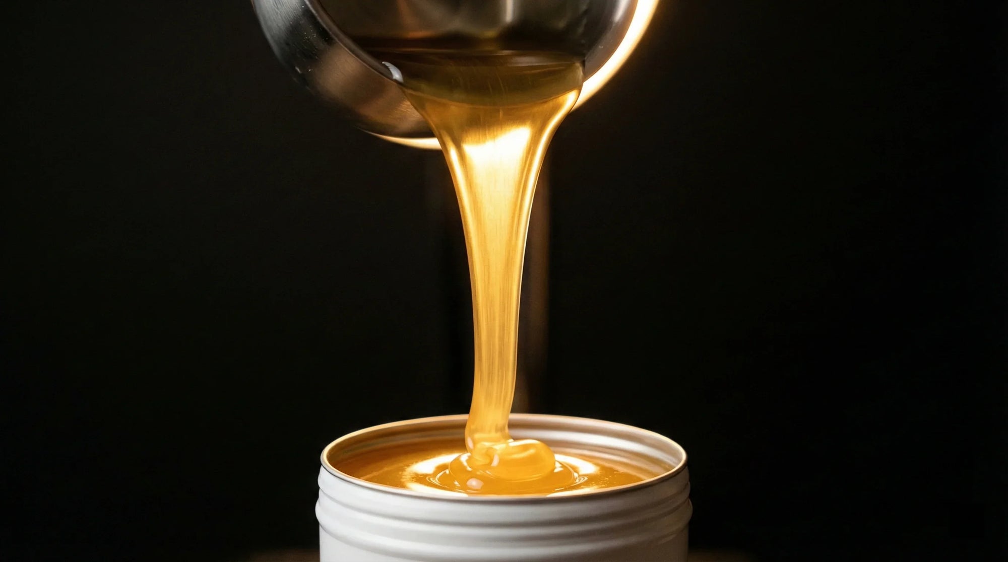 Golden beard balm being poured from a metal ladle into a white tin container against a dark background