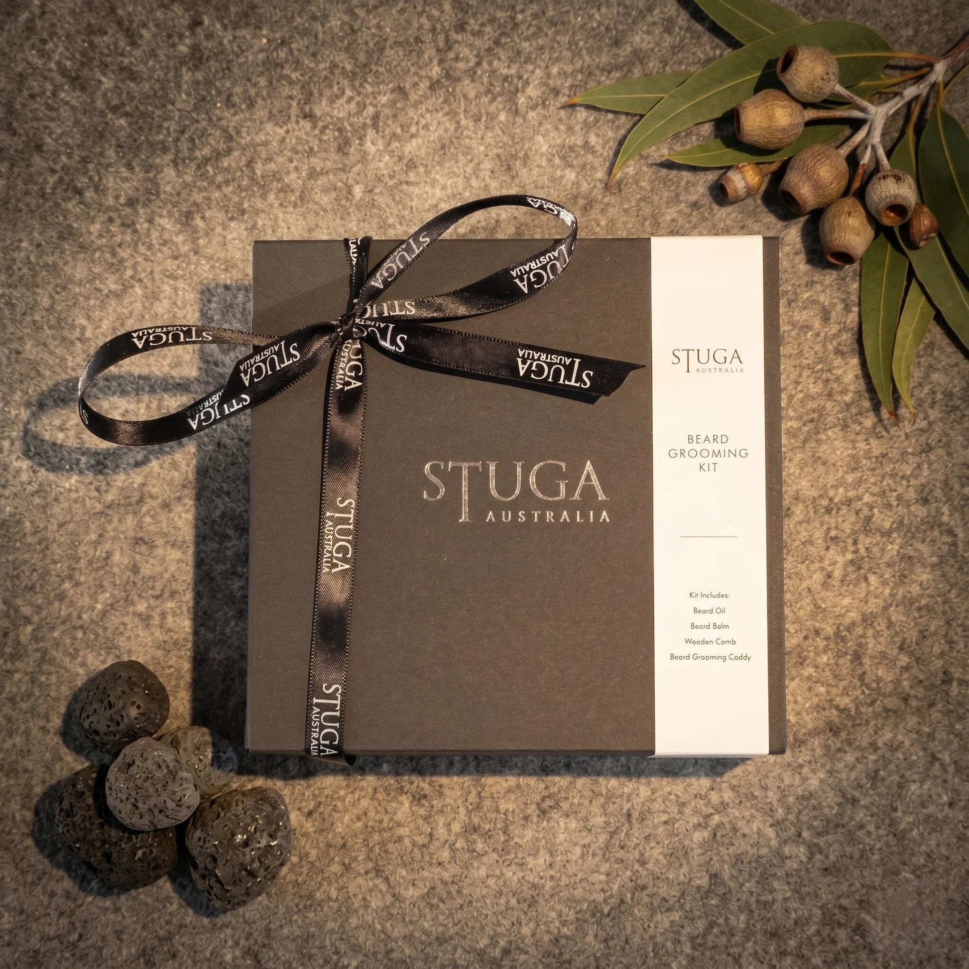 Stuga Beard Grooming Kit in a matte black gift box tied with a Stuga-branded satin ribbon, with eucalyptus leaves and gum nuts on a dark stone background.