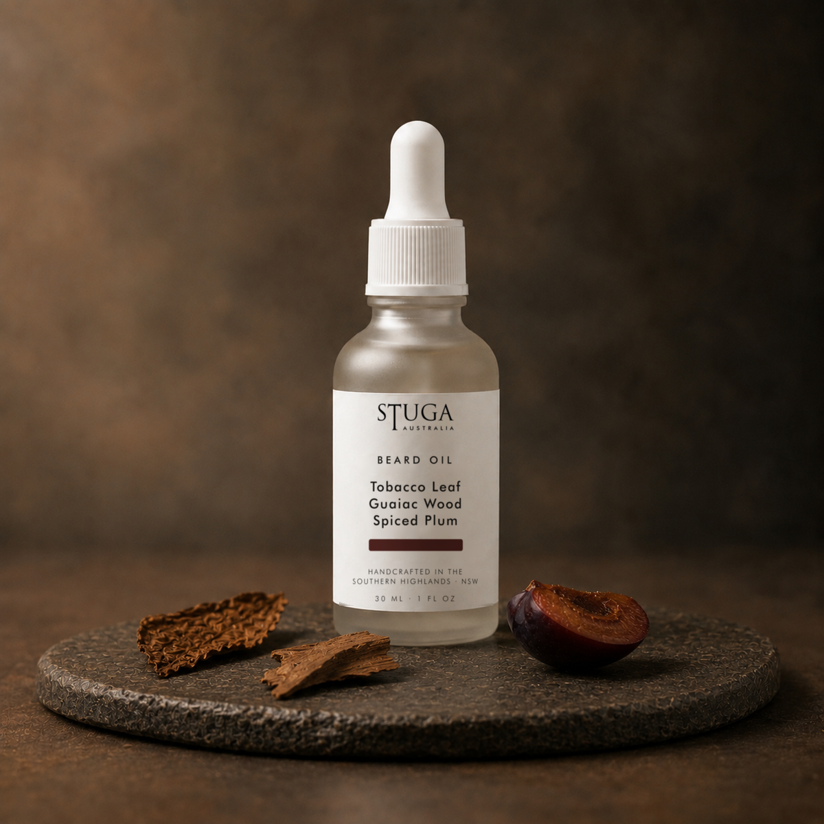 Stuga Beard Oil in Tobacco Leaf, Guaiac Wood and Spiced Plum — frosted glass dropper bottle on a dark stone disc with a curl of cured tobacco leaf, guaiac wood bark and a halved dark plum against a warm brown backdrop. Lightweight beard oil handcrafted in the Southern Highlands, NSW Australia.
