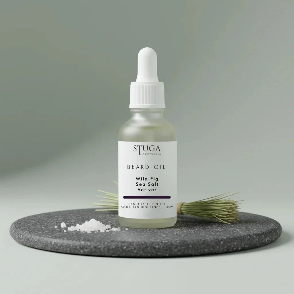 Stuga Beard Oil in Wild Fig, Sea Salt and Vetiver — pale oil in a frosted glass dropper bottle on a dark granite disc with flaked sea salt and a bundle of vetiver root. Made fresh to order with Australian sweet almond, macadamia and apricot kernel oils, handcrafted in the Southern Highlands, NSW Australia.