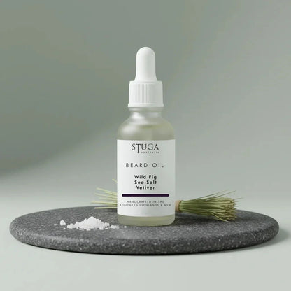 Stuga Beard Oil in Wild Fig, Sea Salt and Vetiver — pale oil in a frosted glass dropper bottle on a dark granite disc with flaked sea salt and a bundle of vetiver root. Made fresh to order with Australian sweet almond, macadamia and apricot kernel oils, handcrafted in the Southern Highlands, NSW Australia.
