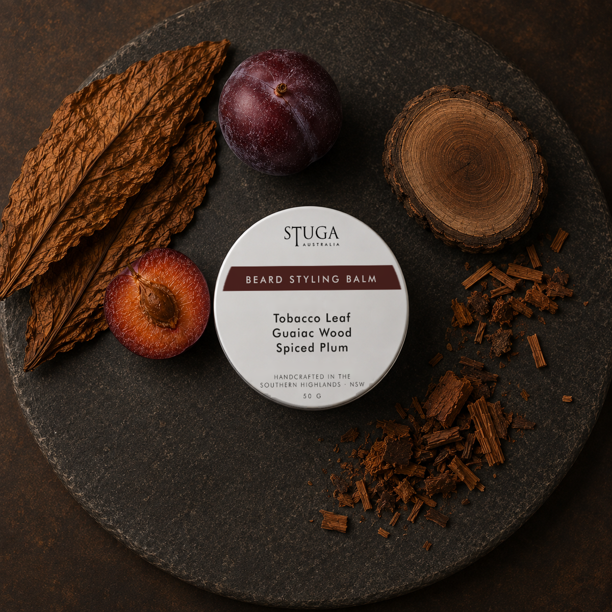 Stuga Beard Styling Balm in Tobacco Leaf, Guaiac Wood and Spiced Plum — 50g white tin on a dark stone slab with cured tobacco leaves, whole and halved dark plums, and a cut round of guaiac wood with shavings. Natural hold beard balm handcrafted in the Southern Highlands, NSW Australia.