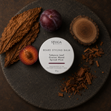 Stuga Beard Styling Balm in Tobacco Leaf, Guaiac Wood and Spiced Plum — 50g white tin on a dark stone slab with cured tobacco leaves, whole and halved dark plums, and a cut round of guaiac wood with shavings. Natural hold beard balm handcrafted in the Southern Highlands, NSW Australia.
