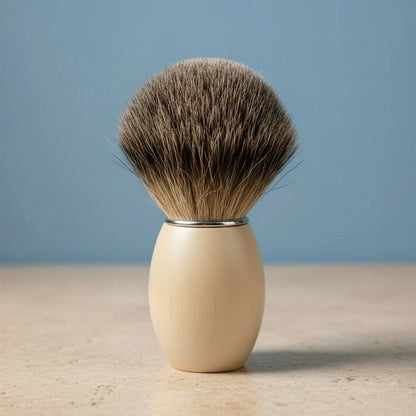 Shaving brush with light beech wood handle and dark 3rd grade badger bristles, standing upright on a light surface with a blu