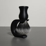 Black shaving brush with synthetic badger hair bristles, resting in a matching black stand on a grey surface.