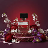 Desire Eau de Parfum 50ml bottle with black cap, surrounded by red peonies, white cherry blossoms, and purple flowers on a cr