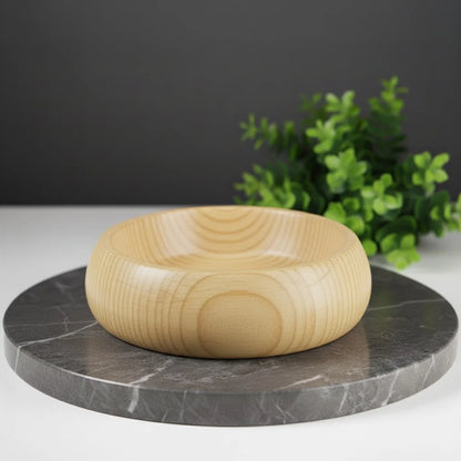 Smooth, light-colored huon pine wood shaving bowl on a dark marble surface with green foliage in the background.