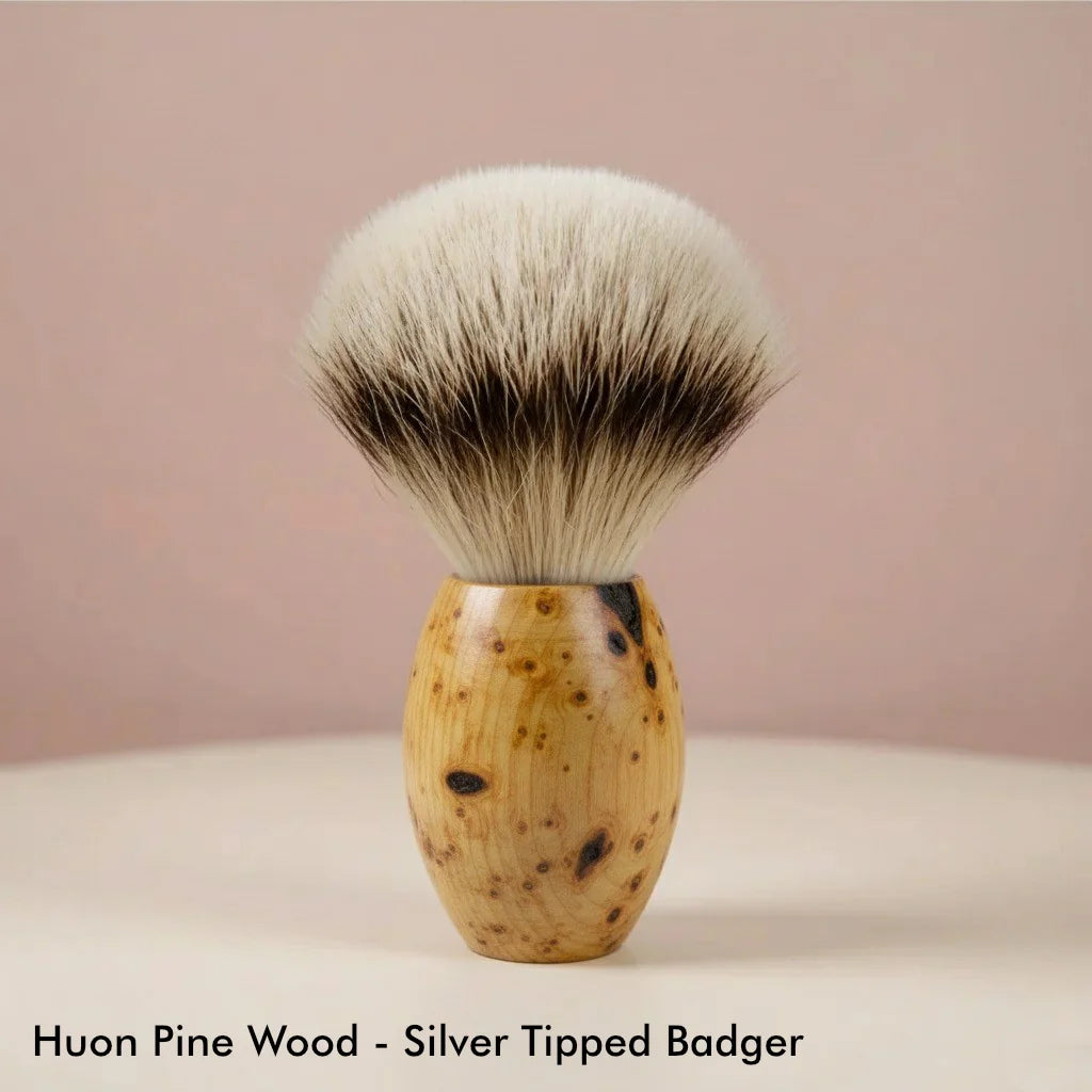 Huon pine wood handle shaving brush with silver tipped badger bristles, light wood grain with dark spots.