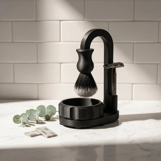 Magic Shaving Kit on a marble bathroom counter — matte black suspension stand with hanging synthetic brush, fluted lathering bowl, safety razor, blade packets and eucalyptus sprig