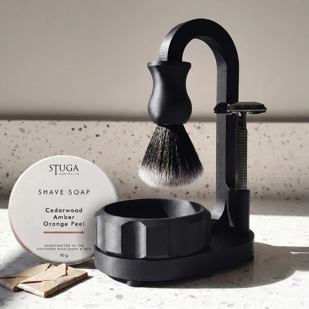 Magic Shaving Kit — matte black polymer stand with suspended synthetic brush, fluted lathering bowl, black safety razor, STUGA shave soap tin and wrapped blades in natural daylight