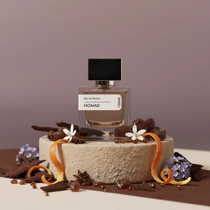 Nomad Eau de Parfum bottle with black cap, surrounded by orange peels, star anise, flowers, and wood chips on a textured ston