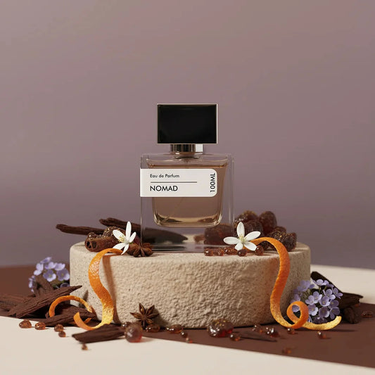Nomad Eau de Parfum bottle with black cap, surrounded by orange peels, star anise, flowers, and wood chips on a textured ston