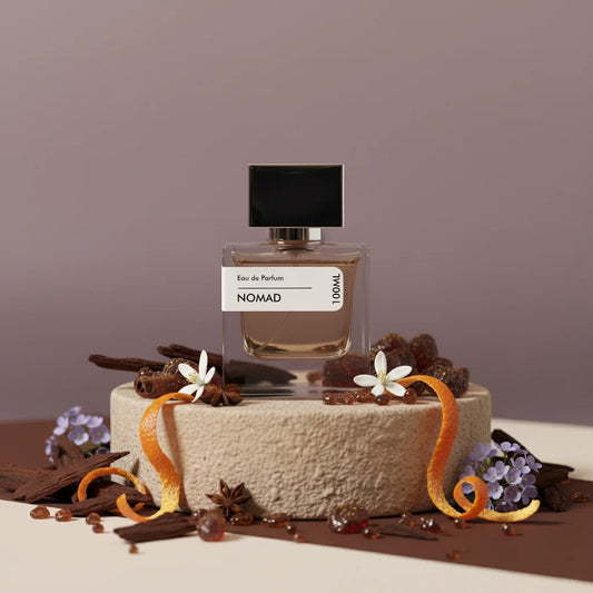 Nomad Eau de Parfum bottle with black cap, surrounded by orange peels, star anise, flowers, and wood chips on a textured ston