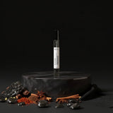 Stuga Obsidian Eau de Parfum 10ml spray bottle on a dark marble pedestal, surrounded by spices and dark stones.