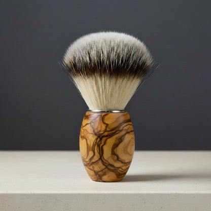 Olive wood shaving brush with synthetic badger-style bristles and a polished metal collar, standing upright on a light surfac