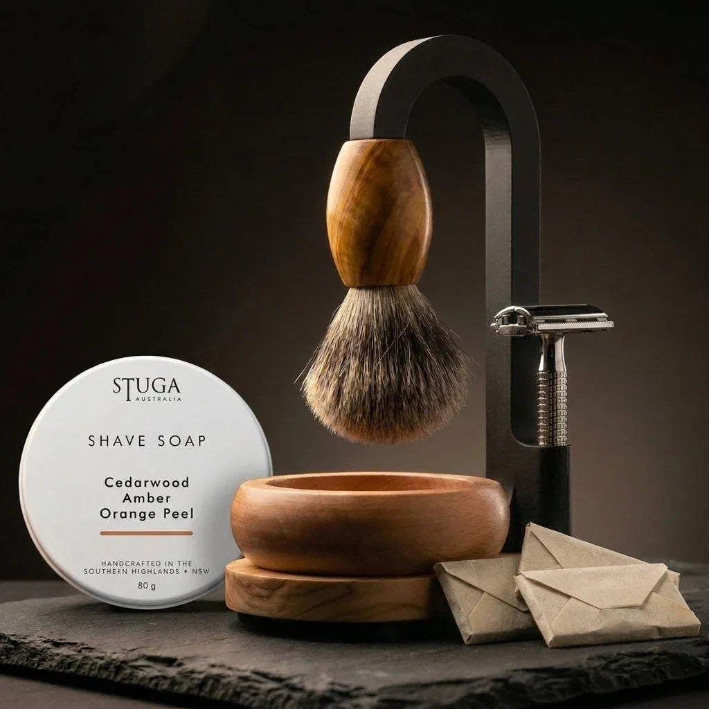 Premium Magic Shaving Kit — badger brush with olive wood handle suspended from black arch stand, STUGA shave soap tin, handturned timber bowl, safety razor and wrapped blades on slate