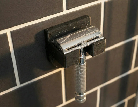 Charcoal black wall-mounted razor holder with chrome safety razor, water droplets visible on dark shower tiles