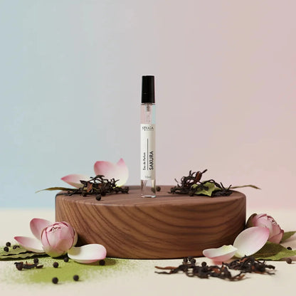 Stuga Sakura Eau de Parfum 10ml rollerball bottle on a wooden stand, surrounded by pink lotus petals and dried tea leaves.