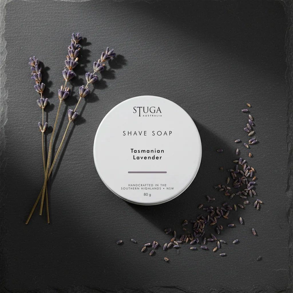 White circular shave soap container with 'STUGA AUSTRALIA' and 'Tasmanian Lavender' text, surrounded by lavender sprigs and loose lavender buds on a dark slate surface.