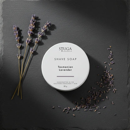 White circular shave soap container with 'STUGA AUSTRALIA' and 'Tasmanian Lavender' text, surrounded by lavender sprigs and loose lavender buds on a dark slate surface.