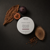 Stuga Artisan Shaving Soap in Tobacco Leaf, Guaiac Wood and Spiced Plum — 80g white tin on dark slate with a cured tobacco leaf, whole and halved dark plums, and a cut round of guaiac wood with shavings. Coconut-oil shave soap handmade in the Southern Highlands, NSW Australia.