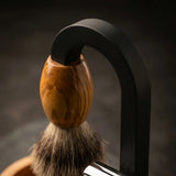 Handcrafted shaving brush with olive wood handle and badger hair bristles on a matte black stand