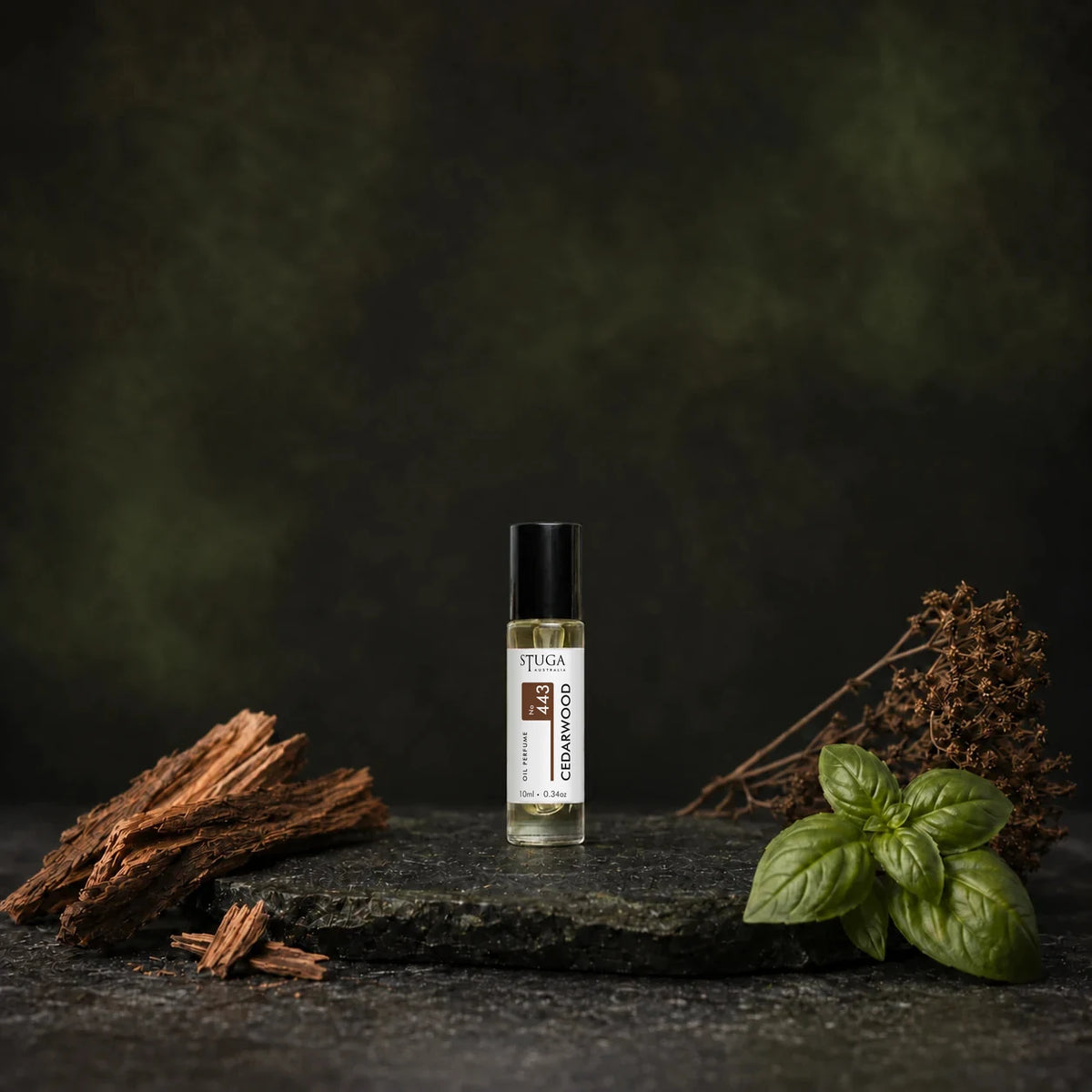 Stuga Cedarwood No. 443 cedarwood oil perfume, 10ml roll-on bottle on dark stone with cedarwood shavings, fresh basil leaves and dried botanicals.