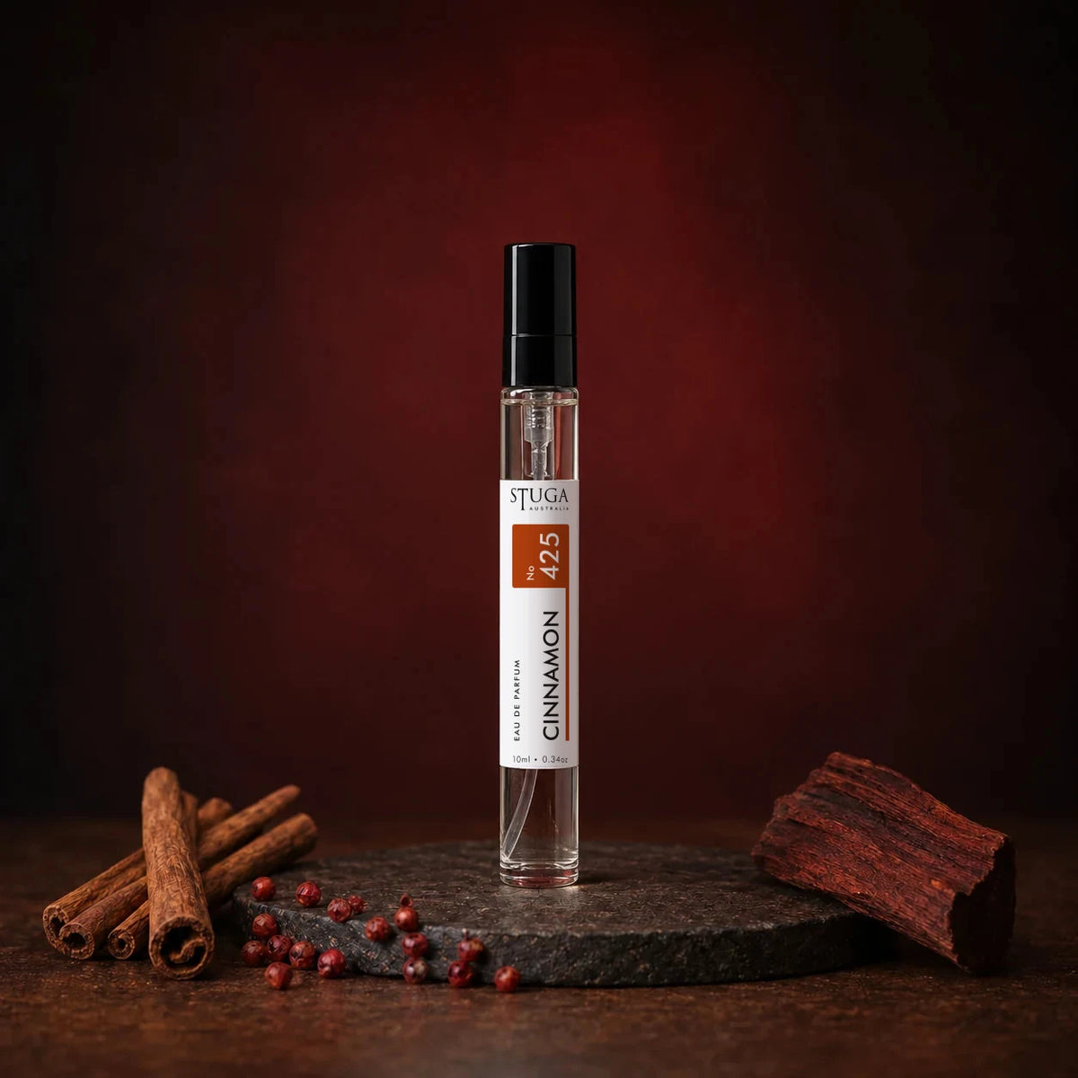 Stuga Cinnamon No. 425 cinnamon perfume, 10ml eau de parfum spray bottle on a dark stone disc with cinnamon sticks, pink peppercorns and mahogany wood against a deep red backdrop.