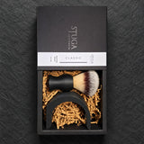 Stuga Classic shaving brush gift box, sliding drawer open with the matte black brush and Wave stand in kraft crinkle paper, on dark slate.
