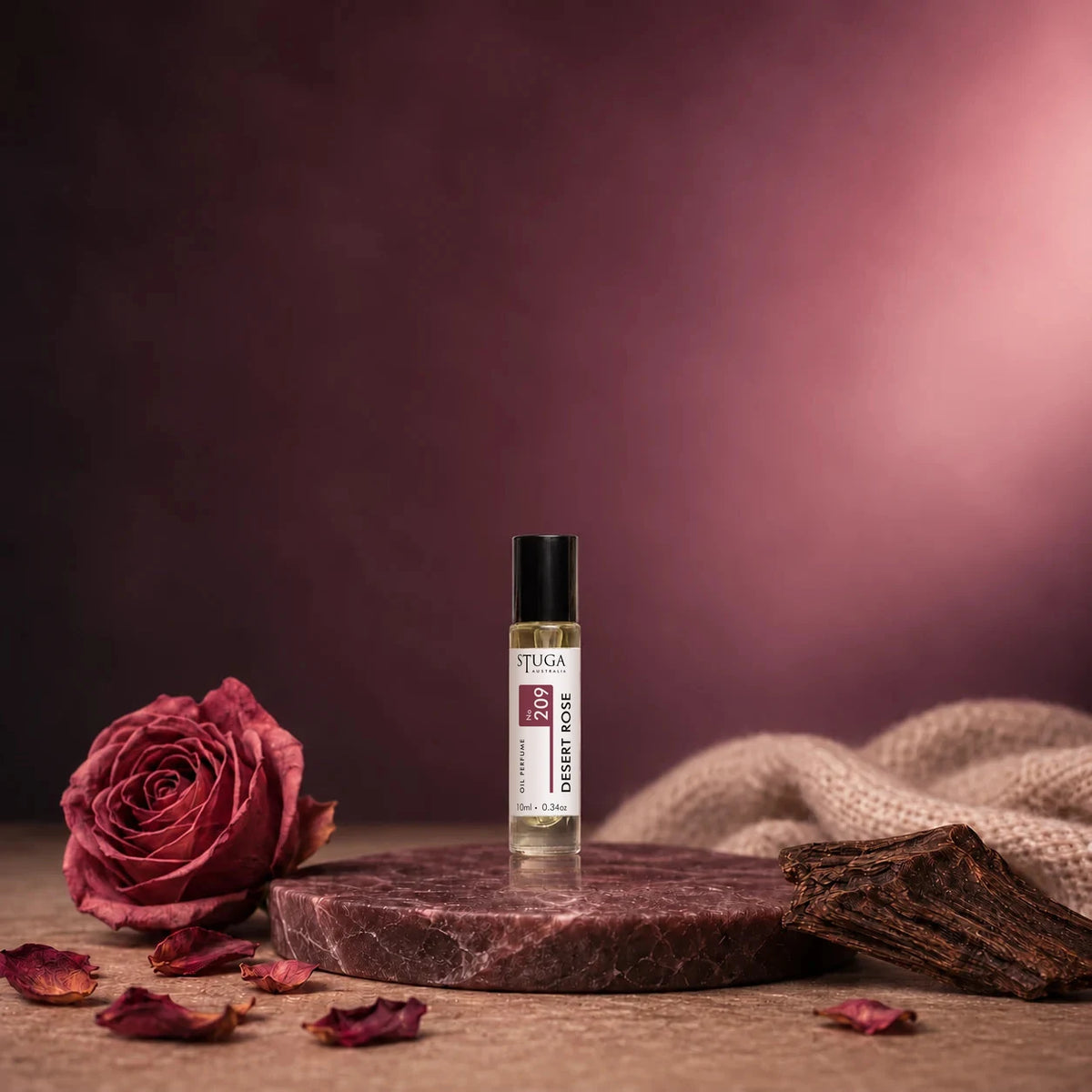 Stuga Desert Rose No. 209 rose oil perfume, 10ml roll-on bottle on a dark marble disc with a dried rose, scattered rose petals, agarwood and cream cashmere.
