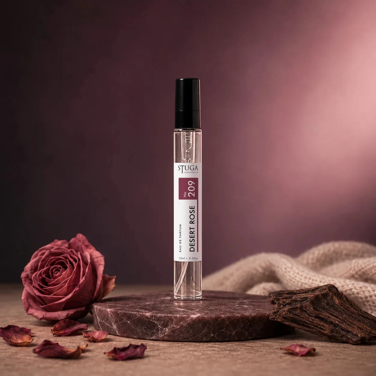 Stuga Desert Rose No. 209 rose perfume, 10ml eau de parfum spray bottle on a dark marble disc with a dried rose, scattered rose petals, agarwood and cream cashmere.