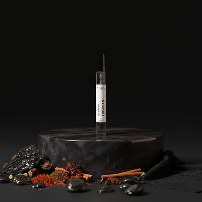 Stuga Obsidian Eau de Parfum 10ml spray bottle on a dark marble pedestal, surrounded by spices and dark stones.