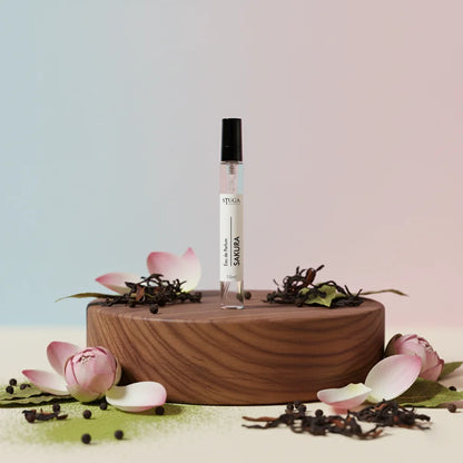 Stuga Sakura Eau de Parfum 10ml rollerball bottle on a wooden stand, surrounded by pink lotus petals and dried tea leaves.