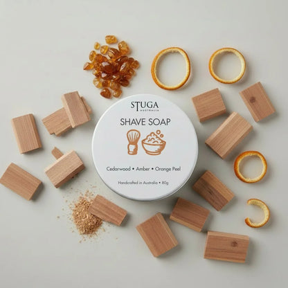 Stuga Australia shave soap tin with cedarwood, amber, and orange peel scents, surrounded by wood blocks and amber pieces.