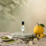 Stuga White Tea No. 203 white tea oil perfume, 10ml roll-on bottle on a pale green ceramic stand with matcha powder, a whole yuzu with peel twist, loose green tea leaves and jasmine blossom.