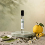 Stuga White Tea No. 203 white tea perfume, 10ml eau de parfum spray bottle on a pale green ceramic stand with matcha powder, a whole yuzu with peel twist, loose green tea leaves and jasmine blossom.