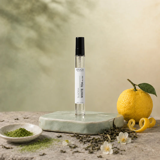 Stuga White Tea No. 203 white tea perfume, 10ml eau de parfum spray bottle on a pale green ceramic stand with matcha powder, a whole yuzu with peel twist, loose green tea leaves and jasmine blossom.