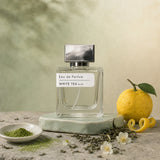 Stuga White Tea No. 203 white tea perfume, 50ml eau de parfum bottle on a pale green ceramic stand with matcha powder, a whole yuzu with peel twist, loose green tea leaves and jasmine blossom.